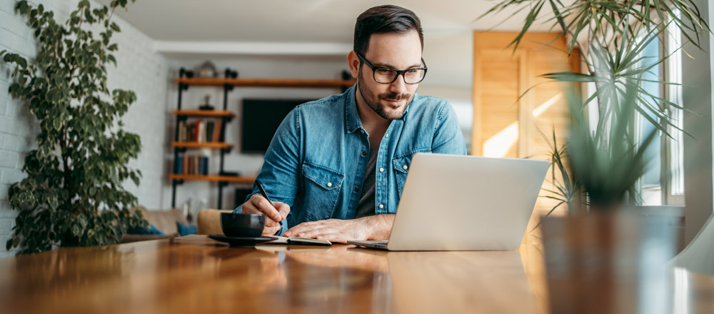 Man wearing glasses using a laptop while writing something on a tablet